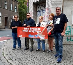 Fünf Politiker:innen stehen auf einer Straße und halten ein Banner hoch auf welchem steht: Den braunen Müll wegfegen.